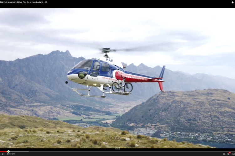 A helicopter hovering above a mountainous landscape, with bicycles attached below it. The scene features rolling hills and distant mountains under a partly cloudy sky, showcasing the natural beauty of New Zealand.