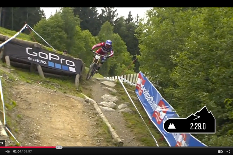 Biker navigating a rocky trail during a mountain biking event, with a GoPro banner in the background and a timer displayed in the corner. Lush greenery surrounds the path.