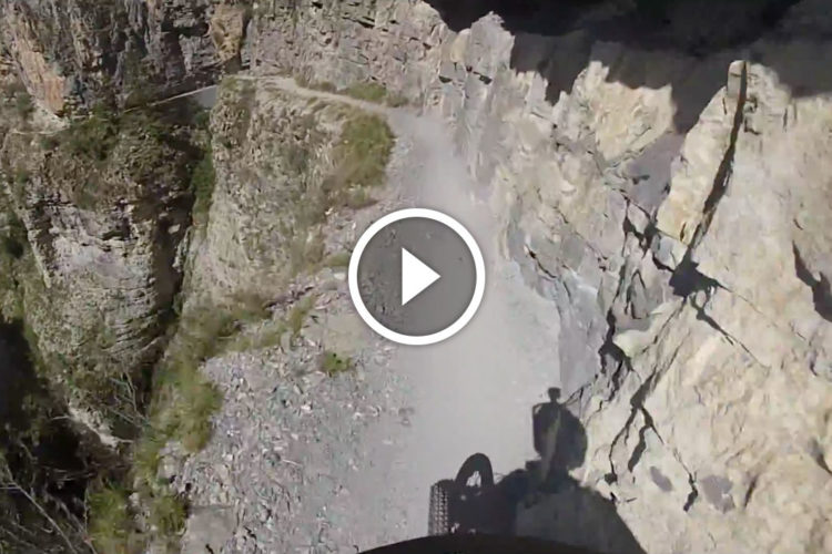 A narrow mountain bike trail along a rocky cliff, with a steep drop to the left and surrounding rock formations. The view shows the edge of the trail, emphasizing the rugged terrain and the shadow of the cyclist. A play button icon indicates that the image is part of a video.