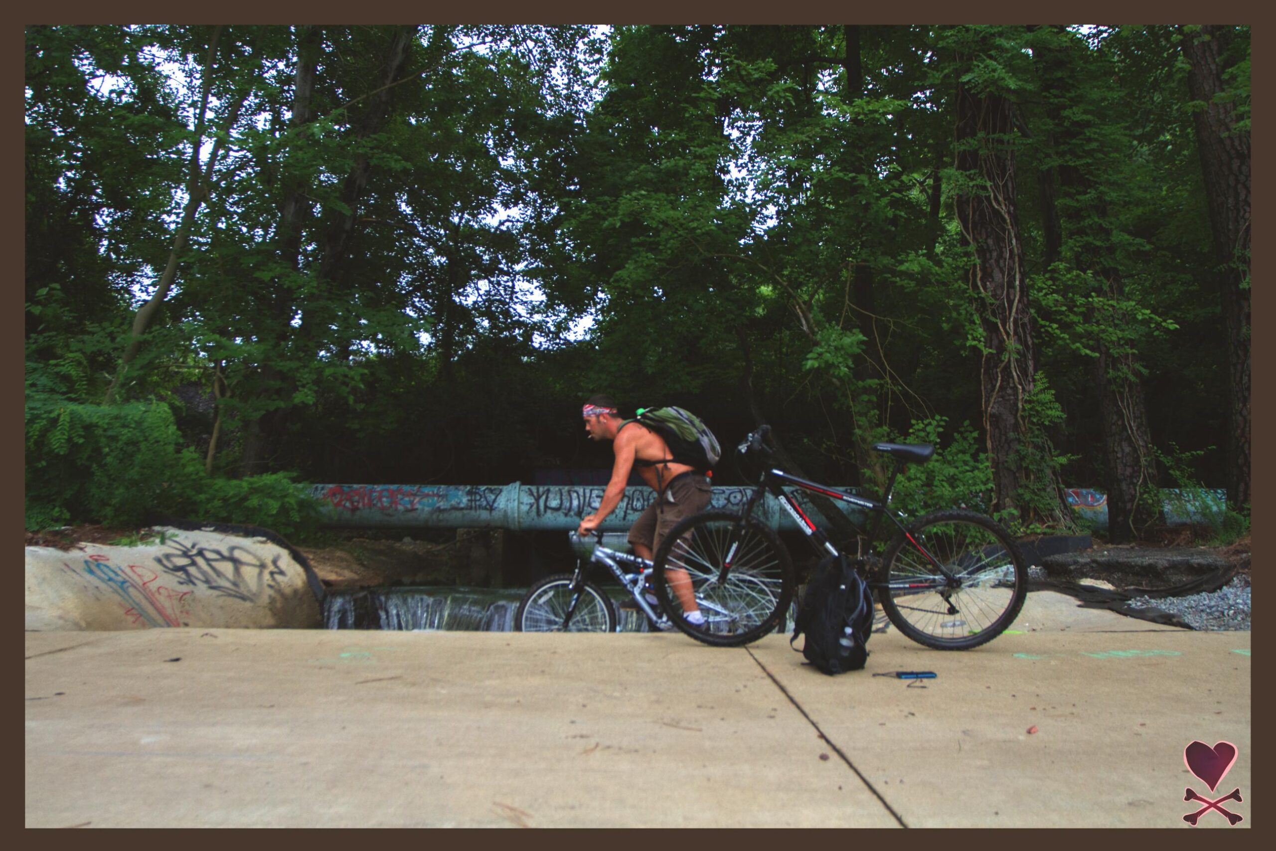A shirtless cyclist in shorts rides his bike near a small waterfall, with a graffiti-covered pipe in the background. Two bicycles are resting on a paved surface, surrounded by lush greenery. The scene is set in a natural environment, suggesting an outdoor recreational area. Lake Maury mountain bike trail.