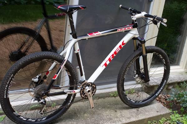 Trek 8000: A white and red Trek 8000 mountain bike is resting against a wall, showcasing its front suspension and knobby tires. The bike features a modern design with a lightweight frame, and it is positioned on a stone surface with a blurred reflection visible in the background.