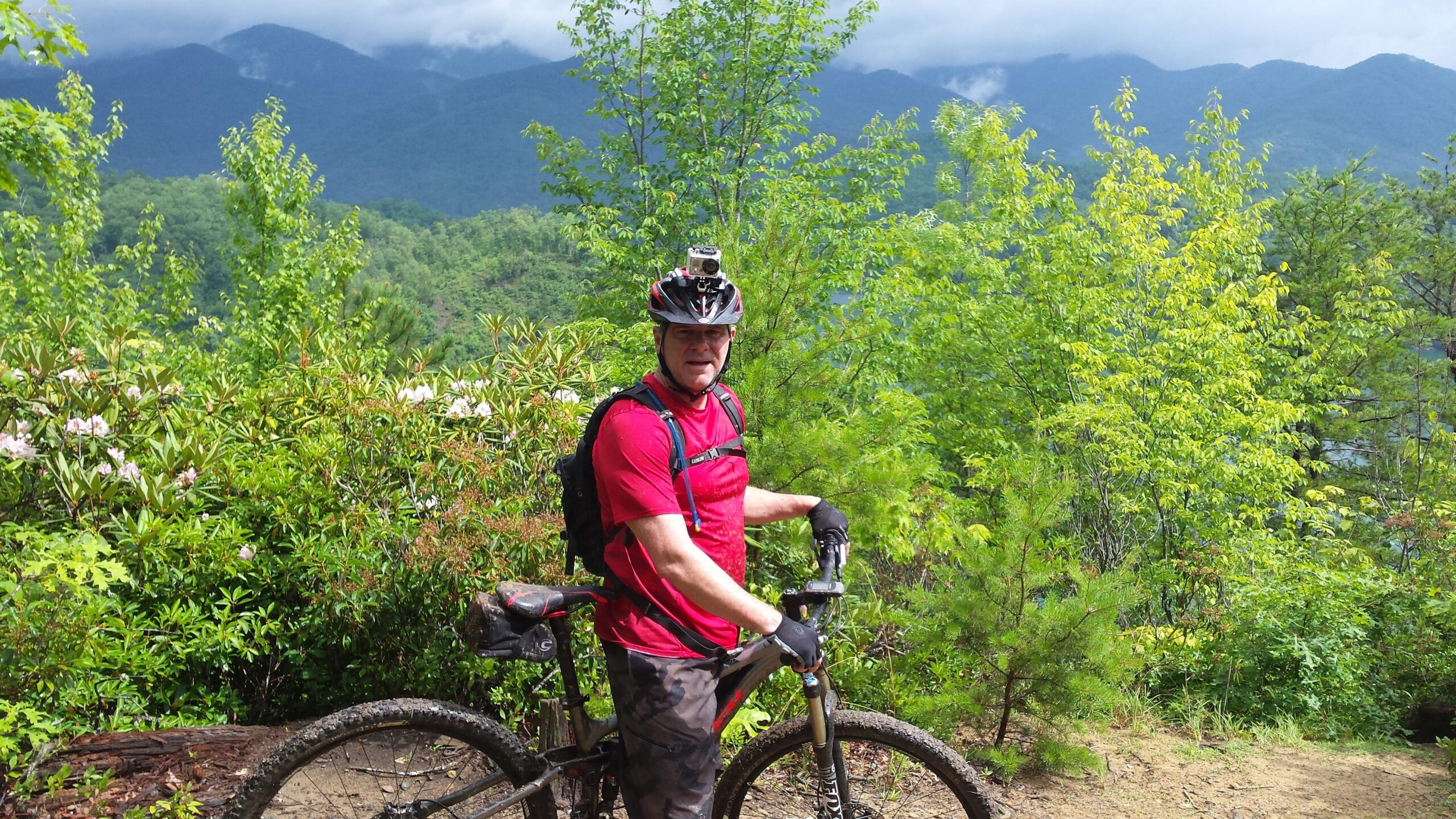 A mountain biker in a red shirt and black gloves stands next to a muddy bike, surrounded by lush greenery and flowering plants, with a backdrop of distant mountains under a partly cloudy sky. Tsali Left Loop mountain bike trail.