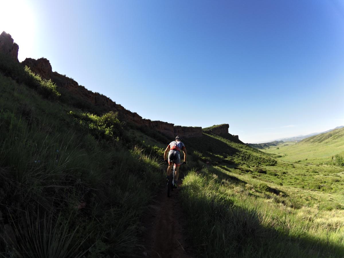 A mountain biker riding along a narrow trail surrounded by lush green hills and rocky outcrops under a clear blue sky. The trail winds through a scenic valley, showcasing a vibrant natural landscape. Blue Sky mountain bike trail.