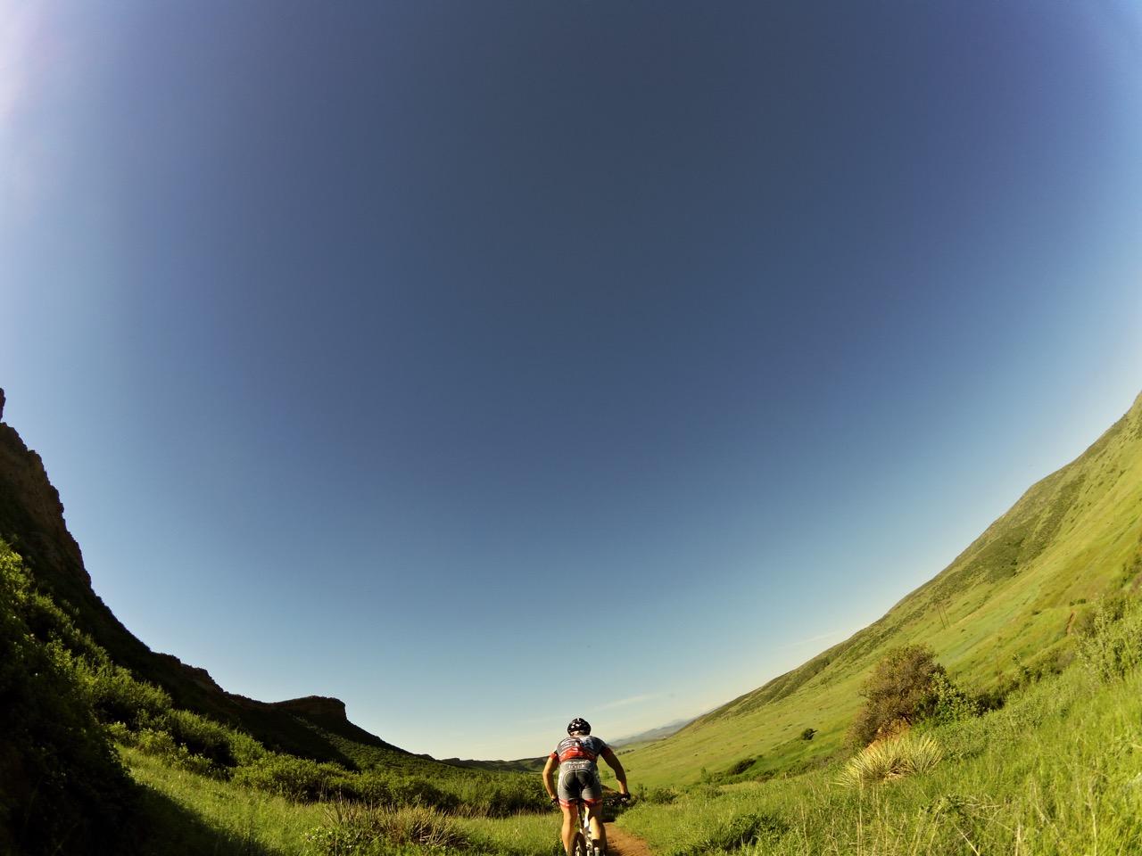A mountain biker riding along a dirt trail in a lush green landscape with rolling hills, under a clear blue sky. Blue Sky mountain bike trail.