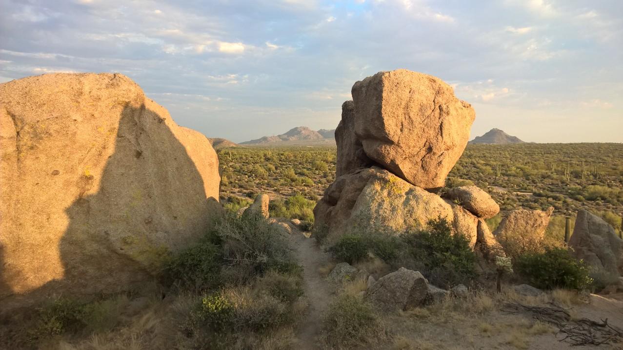 A serene landscape featuring large, rugged boulders in the foreground with a winding path leading into the distance. In the background, rolling hills and distant mountains are visible under a partly cloudy sky, illuminated by soft, golden light. Desert vegetation, including cacti and shrubs, surrounds the rocks, creating a natural and tranquil scene. Pima Road and Dynamite Blvd mountain bike trail.