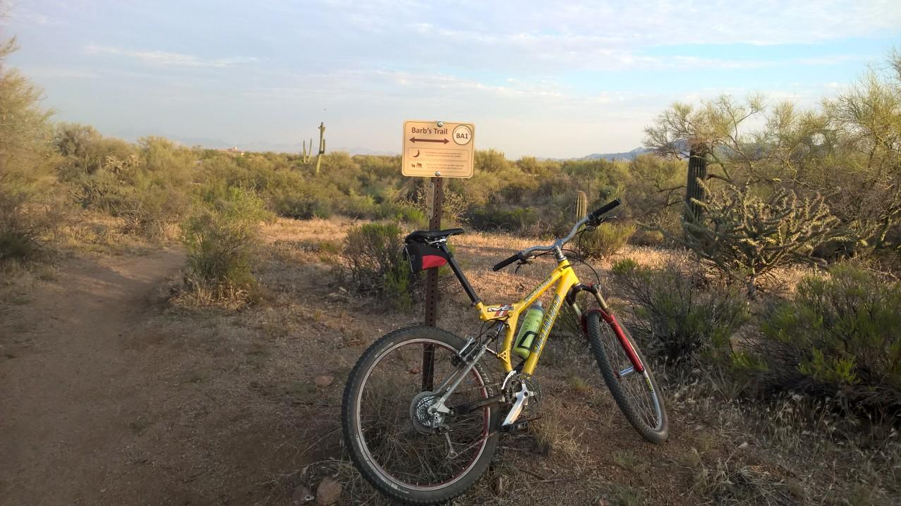A yellow mountain bike rests beside a sign indicating "Barb's Trail" in a desert landscape filled with shrubs and cacti. The trail is surrounded by rolling hills and a clear sky, suggesting a peaceful outdoor environment ideal for biking. Pima Road and Dynamite Blvd mountain bike trail.