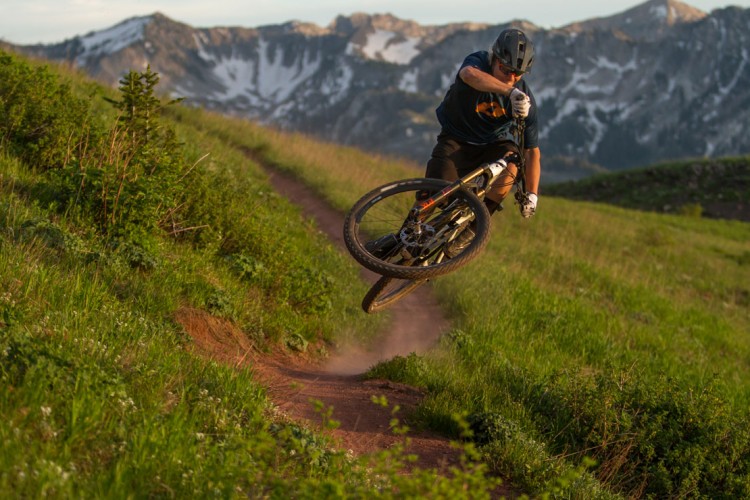 A mountain biker performing a jump on a dirt trail surrounded by lush green grass and distant snow-capped mountains during sunset. Dust is kicked up from the tires as the rider showcases a dynamic biking maneuver.
