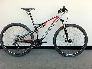 Specialized Epic Comp 29er: Mountain bike with a black and red frame, front suspension, and wide tires, positioned against a white wall on a carpeted floor.
