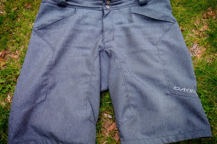 A pair of gray outdoor shorts laid flat on grass, featuring a button and zipper closure, front pockets, and the brand name "DAKINE" displayed on the lower right side.
