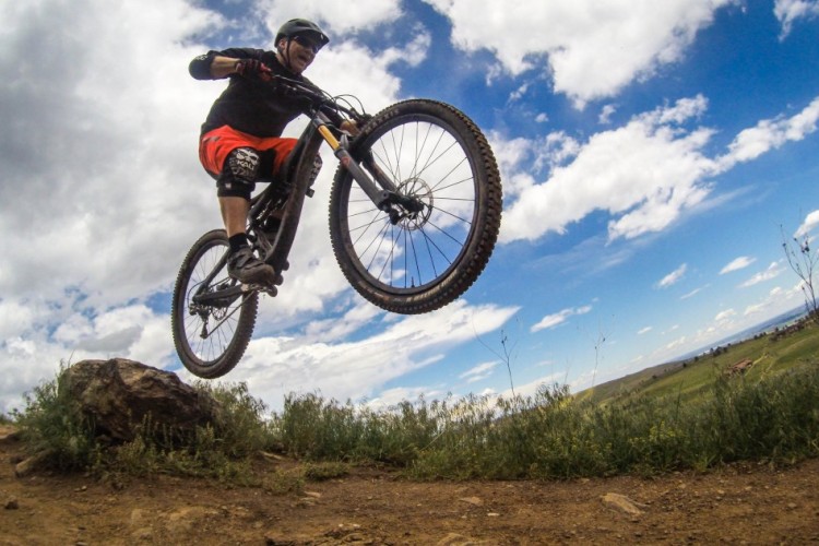 A mountain biker in mid-air as they jump off a rocky terrain, surrounded by green grass and a scenic landscape under a blue sky with clouds.