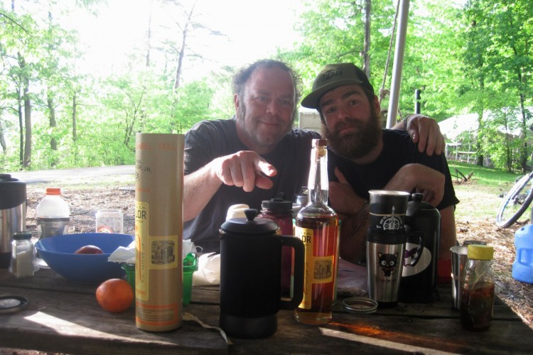 Two men sit together at a picnic table outdoors, surrounded by various items including bottles, mugs, and food. One man points playfully at the camera, while the other smiles and rests his arm on his companion's shoulder. The scene is set in a lush green environment with trees in the background, suggesting a relaxed outdoor gathering or camping experience.