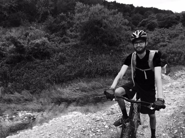 A man wearing a helmet and glasses smiles while riding a mountain bike on a gravel path surrounded by greenery. The image is in black and white, highlighting the outdoor setting and his casual biking attire. Goodwater Trail mountain bike trail.
