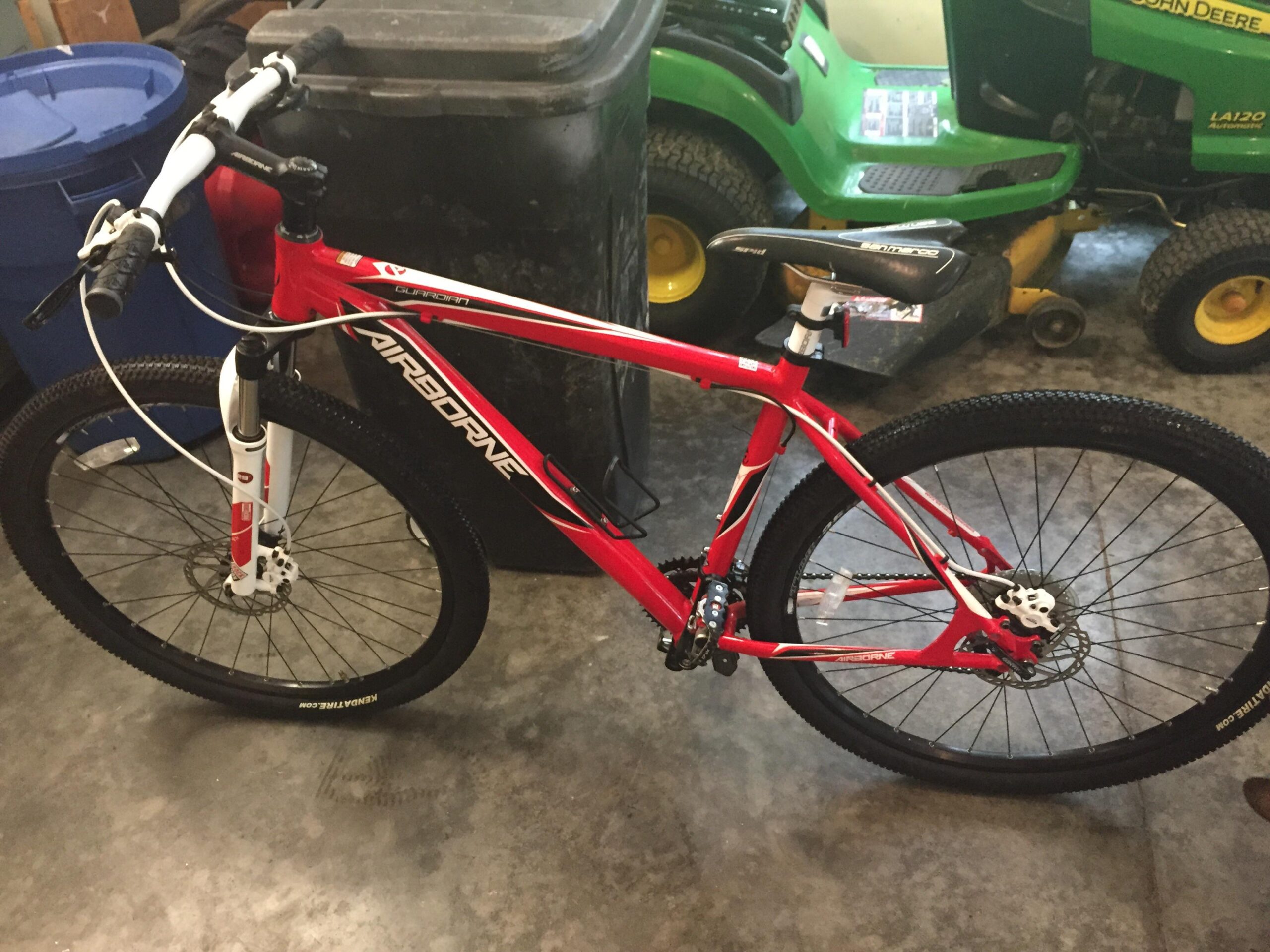 Airborne Guardian: A red mountain bike with white accents, featuring a front suspension fork and disc brakes, positioned in a garage setting with recycling bins and a lawn tractor in the background.