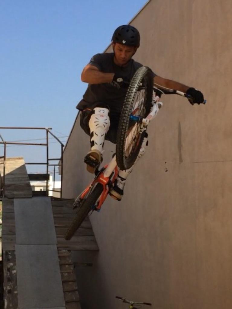 Cannondale Jekyll 3: A mountain biker performing a mid-air jump over a wooden ramp, showcasing a dynamic trick. The rider is wearing a black helmet and protective gear, with the bike partially airborne and one wheel raised high. In the background, there are construction elements visible, hinting at a biking park or training area.