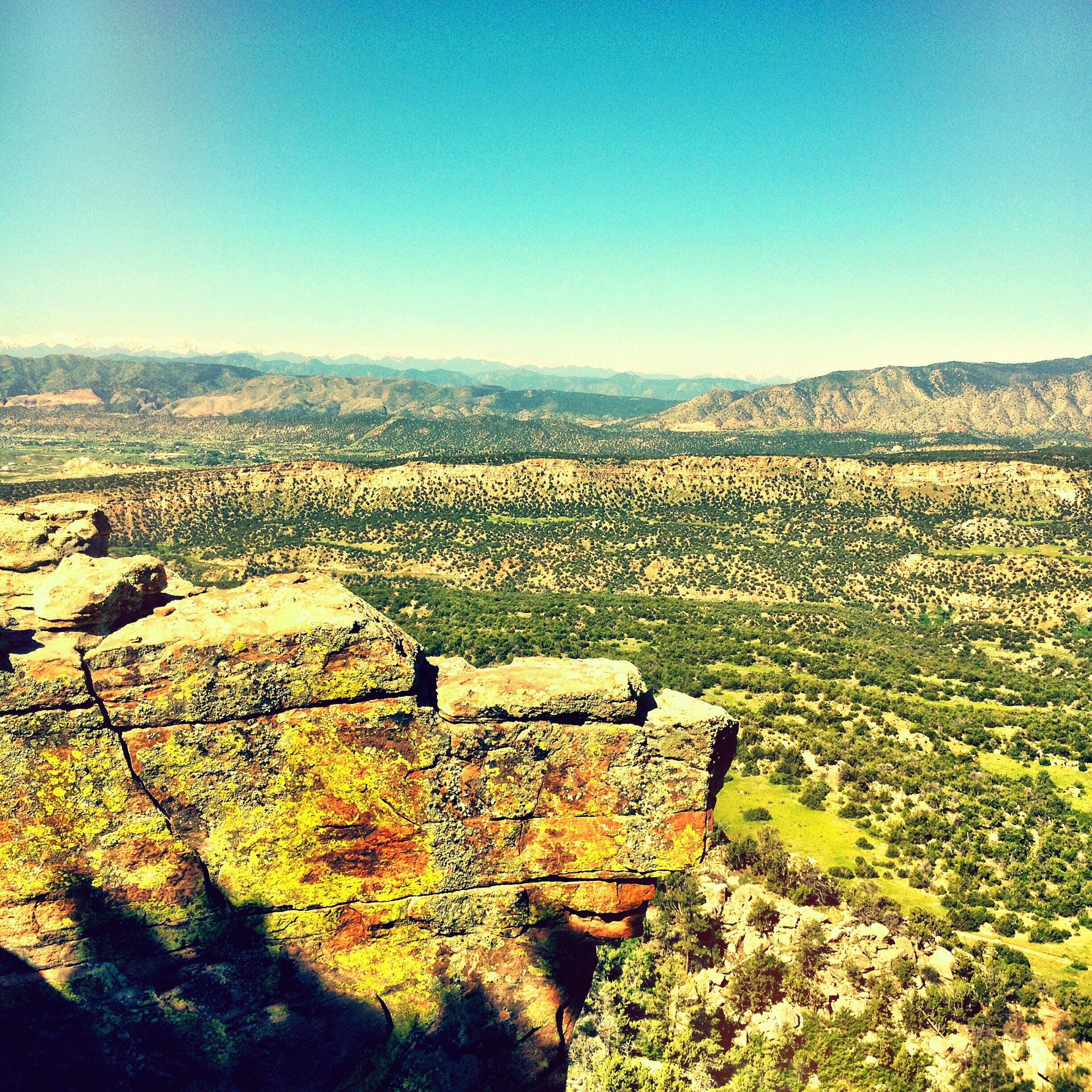 A scenic landscape view from a rocky cliff, showcasing rolling hills and valleys dotted with greenery under a clear blue sky. Distinctive layered rock formations in the foreground contrast with the expansive mountains in the background. Oil Well Flats mountain bike trail.