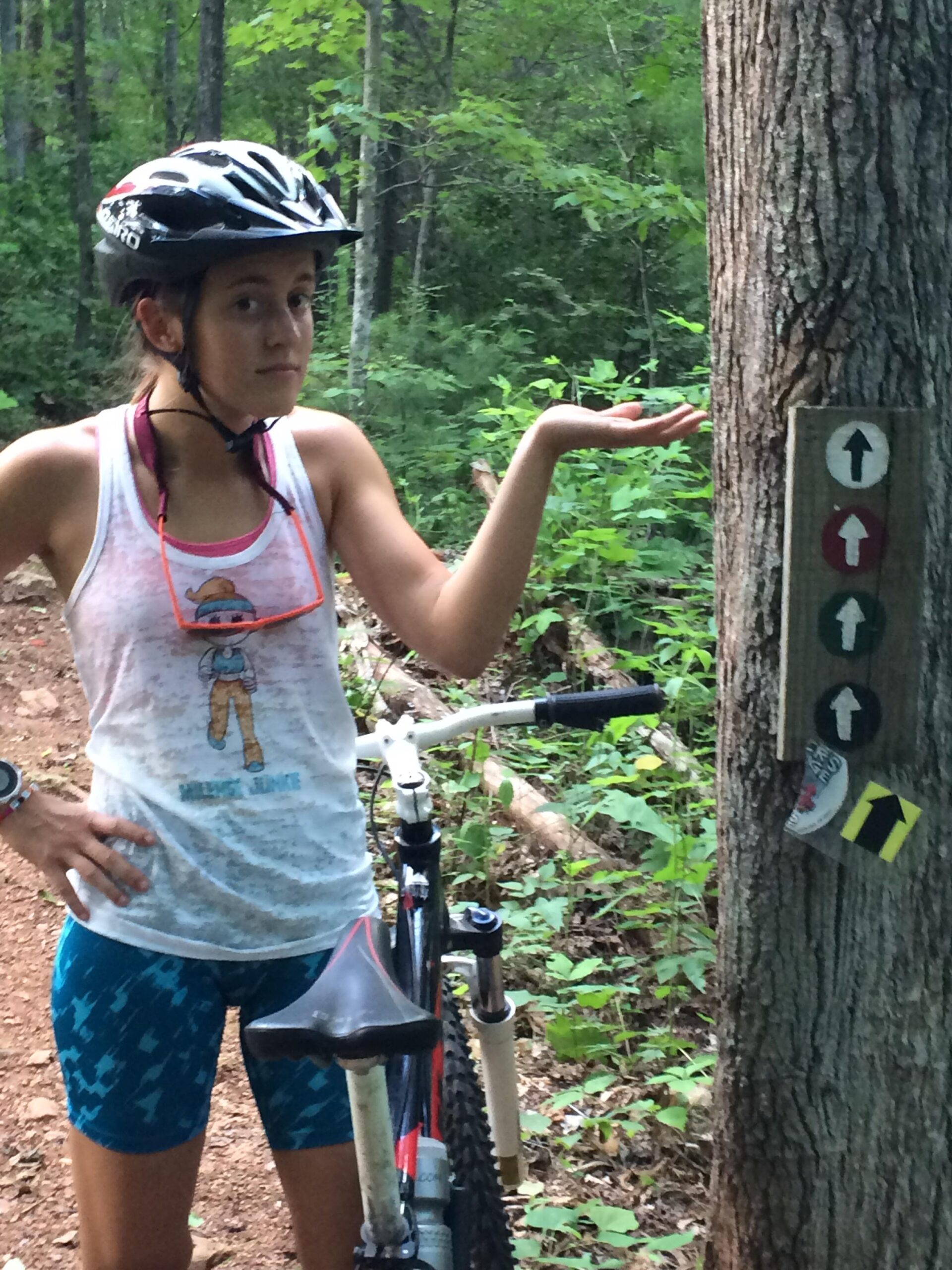 A young person wearing a bike helmet and athletic clothing stands next to a mountain bike, looking puzzled and gesturing with one hand. They are surrounded by dense greenery in a forest setting, with a trail sign mounted on a nearby tree that features directional arrows and markings. Woolwine Trails [Shiners Revenge] mountain bike trail.