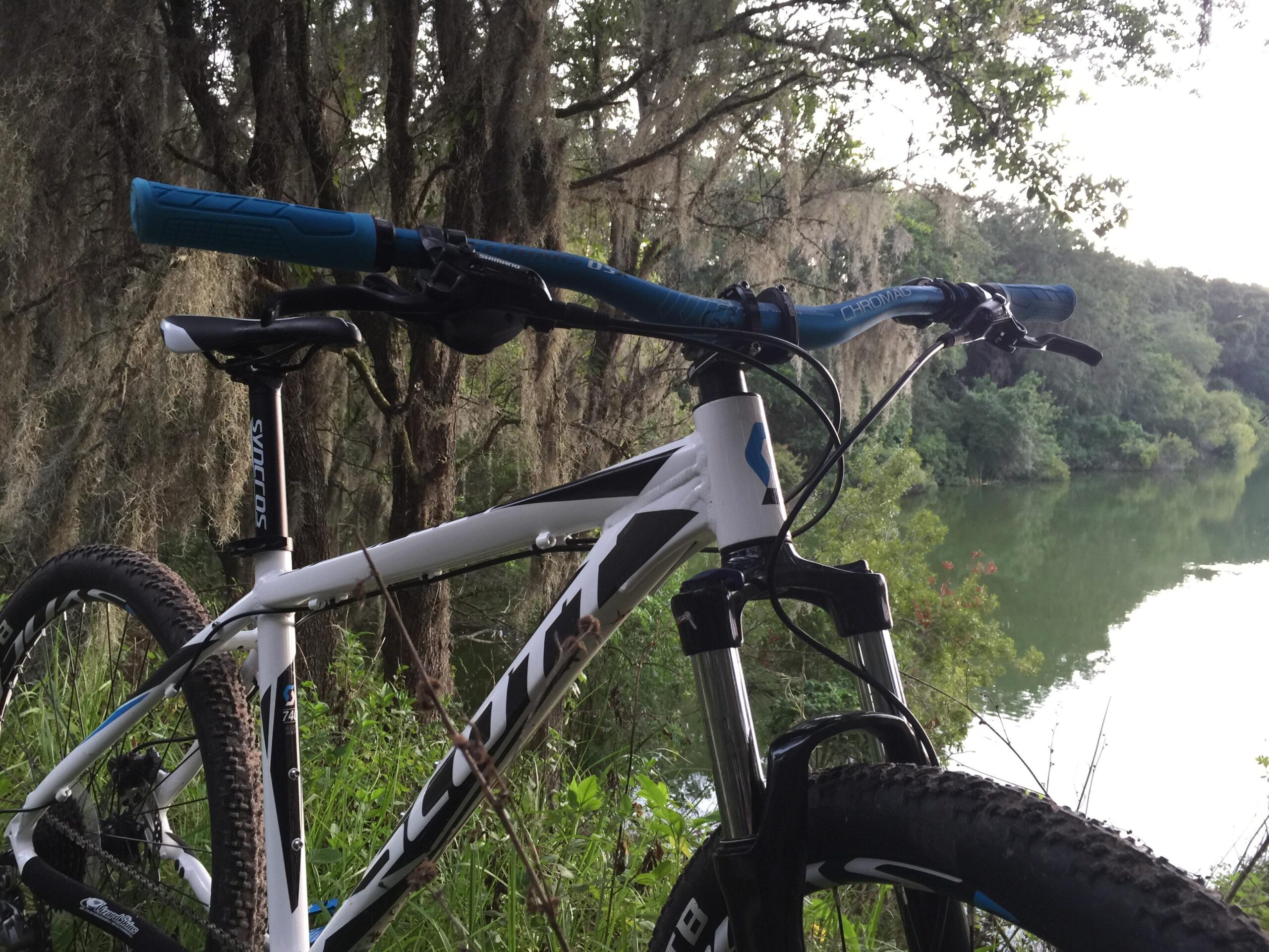 Mountain bike positioned near a calm river surrounded by lush greenery and trees, with the sun setting in the background. The bike features a white frame with blue accents, and the handlebars and seat are clearly visible. Balm Boyette Scrub Preserve mountain bike trail.