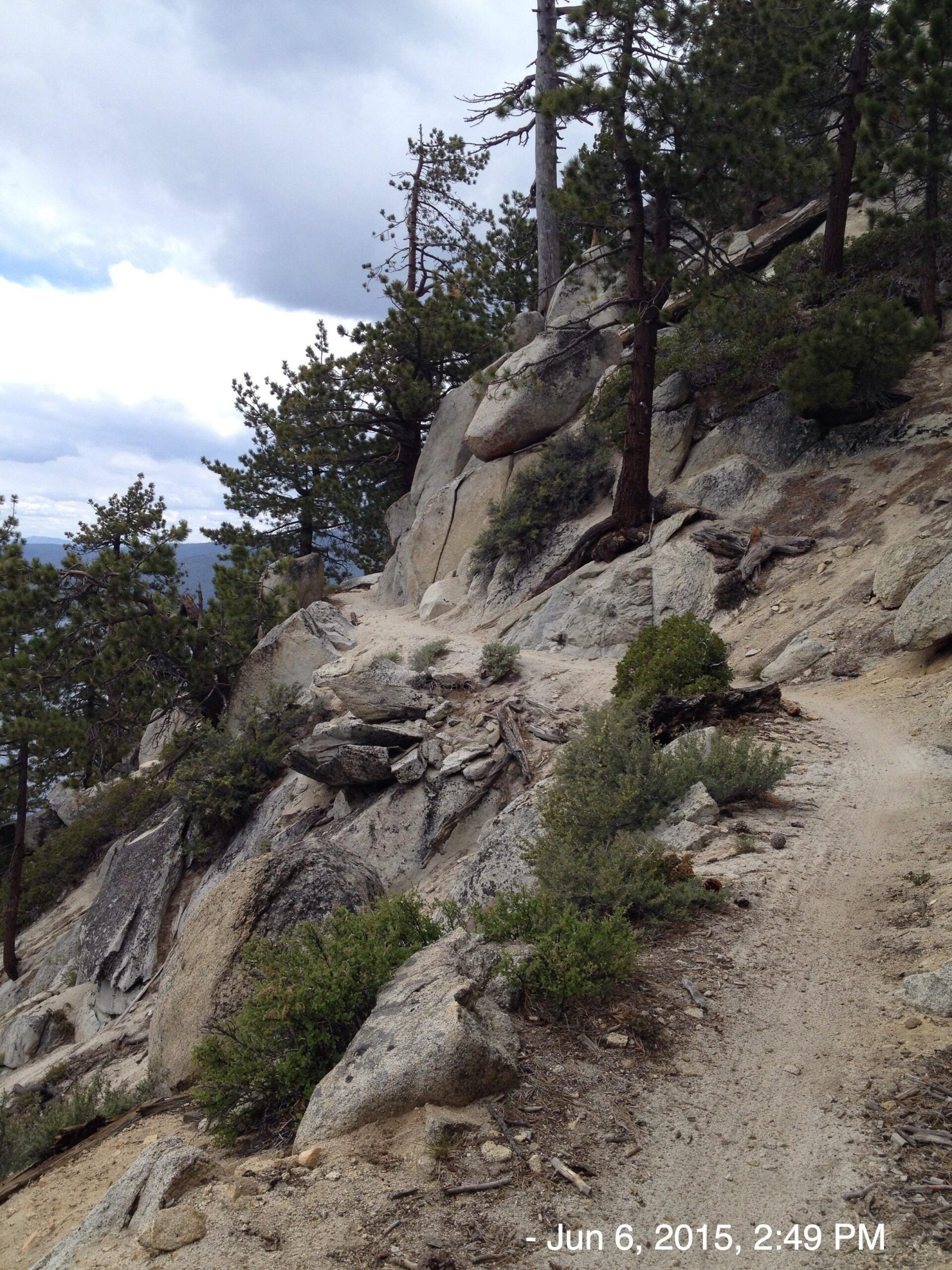 A rocky hiking path winds along a hillside, bordered by trees and shrubs. The sky is partly cloudy, creating a dramatic backdrop for the natural landscape. Tahoe Rim Trail: Tahoe Meadows to Tunnel Creek Road / Flume Trail mountain bike trail.