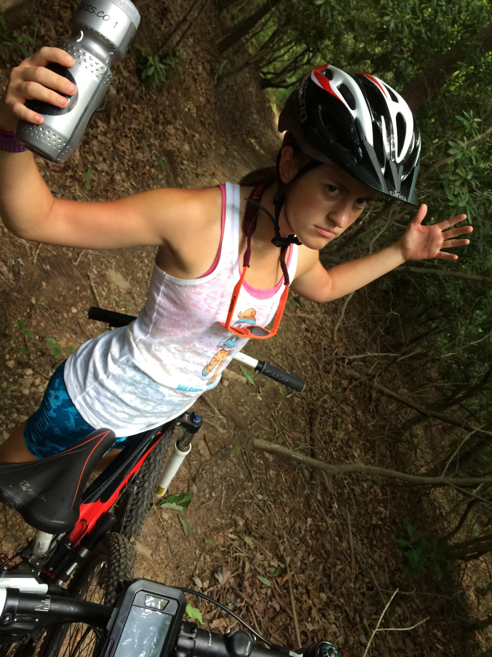 A person wearing a bicycle helmet is standing beside a mountain bike on a dirt trail surrounded by trees. They are holding a water bottle in one hand while extending their other arm outward, appearing to communicate or express a point about the bike ride. The person is dressed in a light tank top and vibrant shorts, showcasing an active outdoor lifestyle. Woolwine Trails [Shiners Revenge] mountain bike trail.