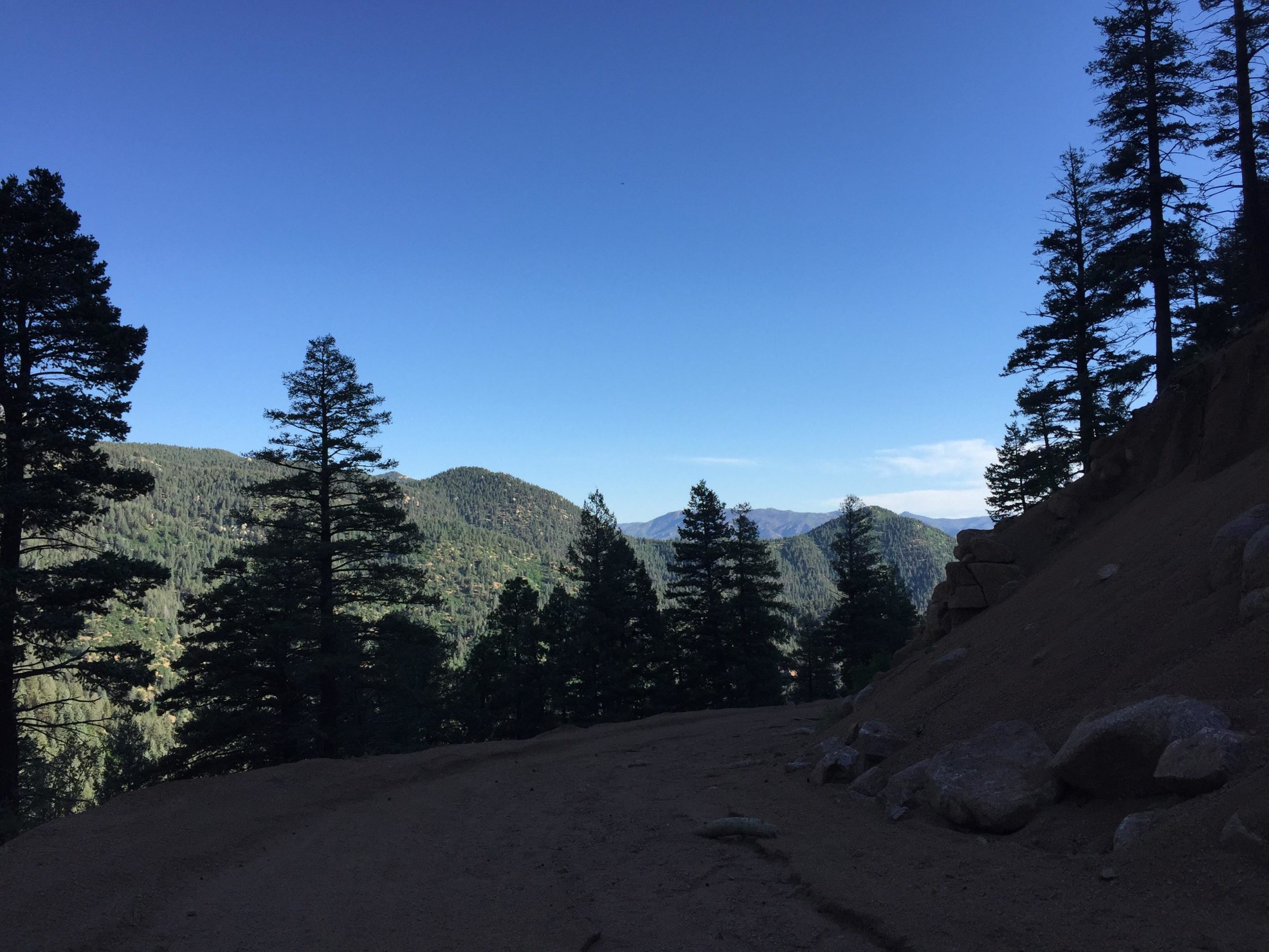 A panoramic view of a mountainous landscape featuring tall evergreen trees, a clear blue sky, and rolling hills in the distance. The foreground includes a dirt path with rocky terrain, while the background showcases layers of lush green mountains under bright sunlight. Stratton Open Space / The Chutes mountain bike trail.