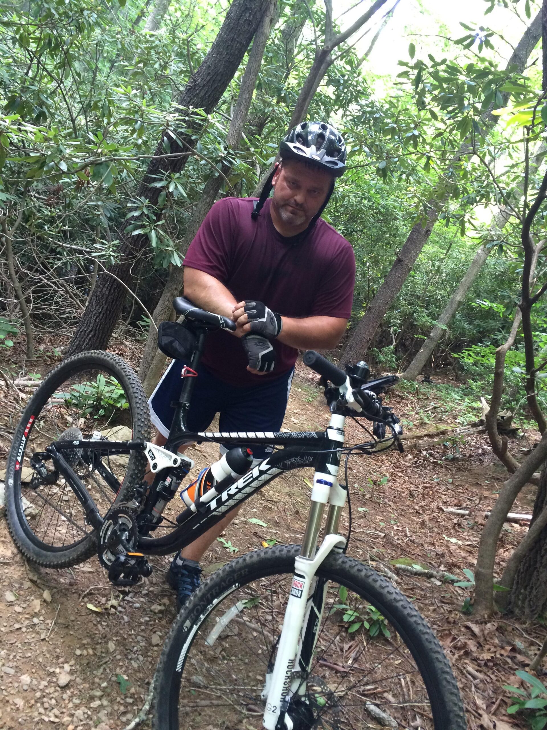 A man in a bicycle helmet stands next to a mountain bike on a dirt trail surrounded by trees and foliage. He is wearing a maroon t-shirt, black fingerless gloves, and shorts, and appears to be adjusting his watch or gear. The scene captures a moment during a mountain biking adventure in a wooded area. Woolwine Trails [Shiners Revenge] mountain bike trail.