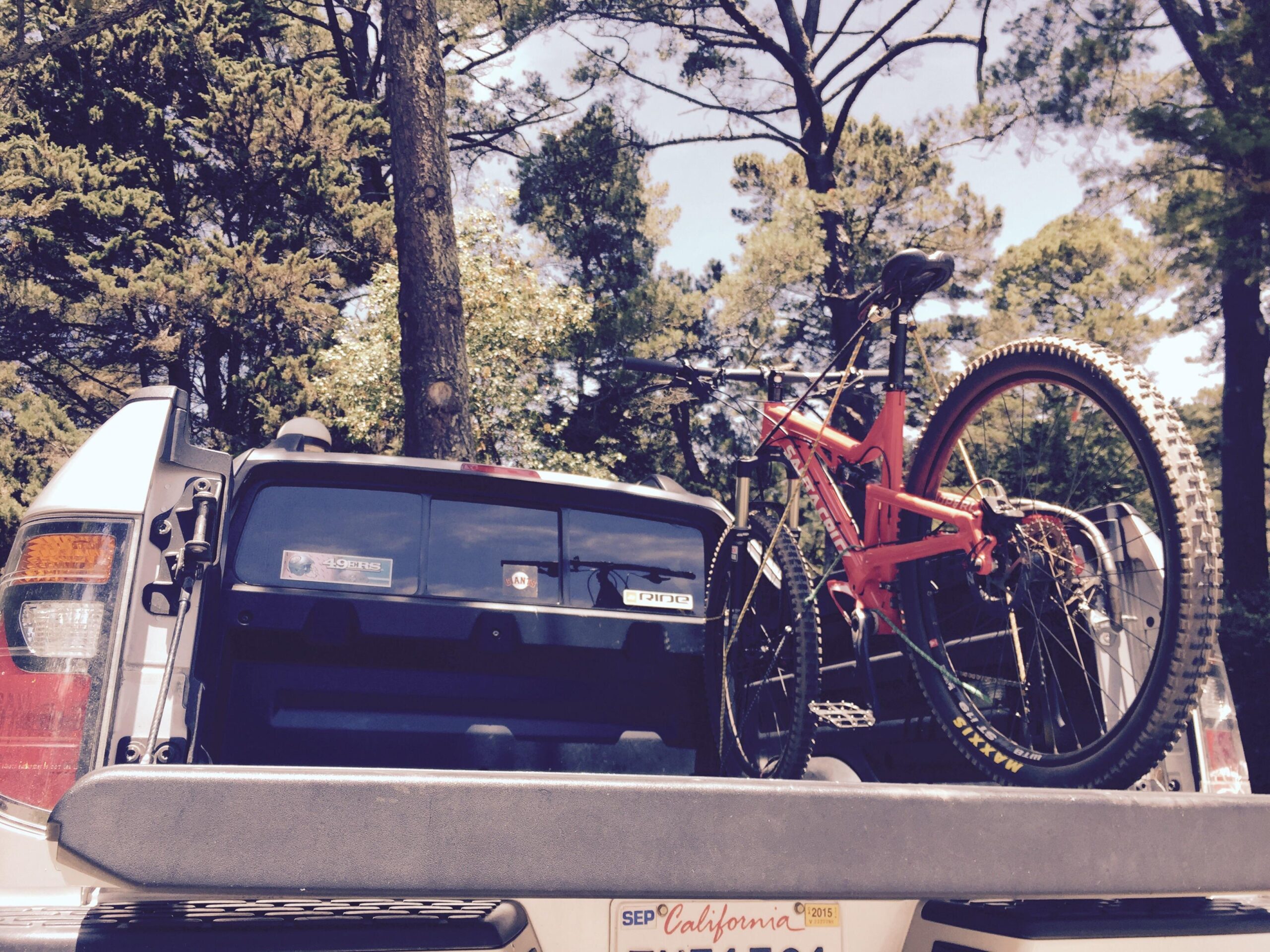 Santa Cruz Superlight: A red mountain bike is securely fastened in the bed of a pickup truck, surrounded by tall green trees. The truck's tailgate is partially open, revealing stickers on the back window and a license plate. The scene is bright and sunny, suggesting an outdoor adventure.