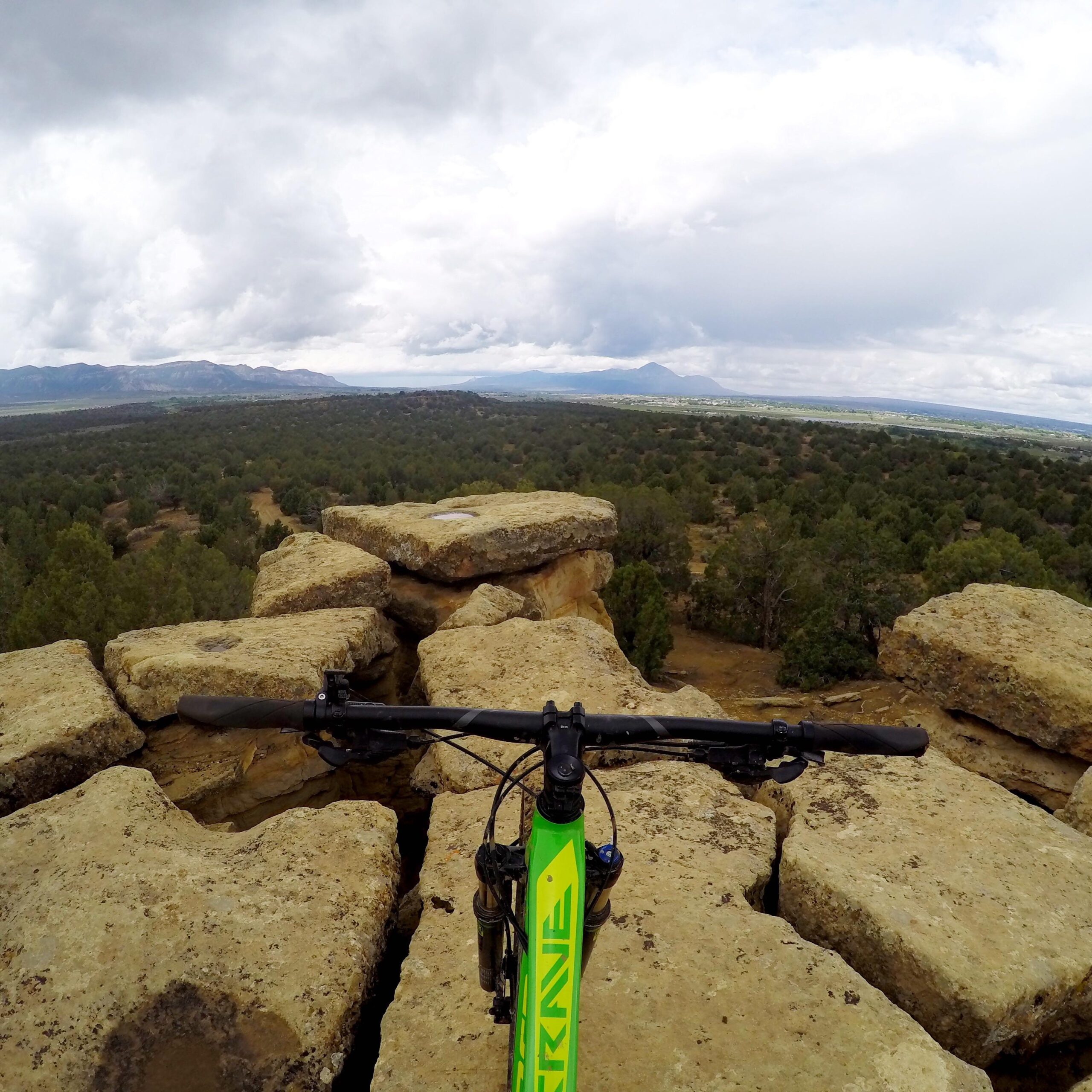 A mountain bike resting on rocky terrain with a panoramic view of green landscape, trees, and mountains under a cloudy sky. Phil's World mountain bike trail.
