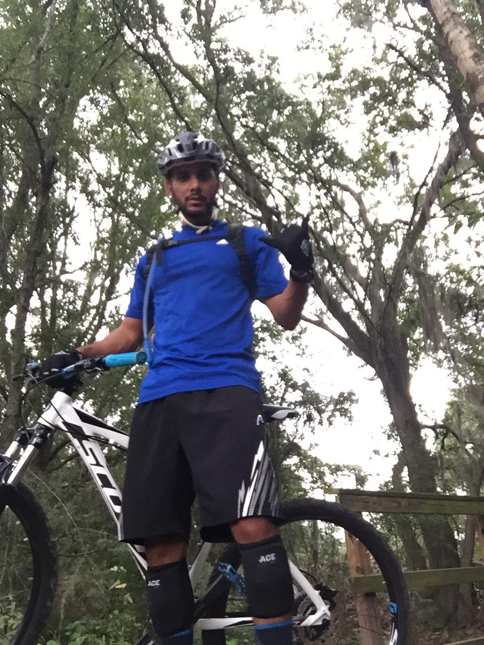 A cyclist wearing a blue shirt, black shorts, and protective gear stands beside a mountain bike in a wooded area, giving a thumbs-up gesture. Balm Boyette Scrub Preserve mountain bike trail.