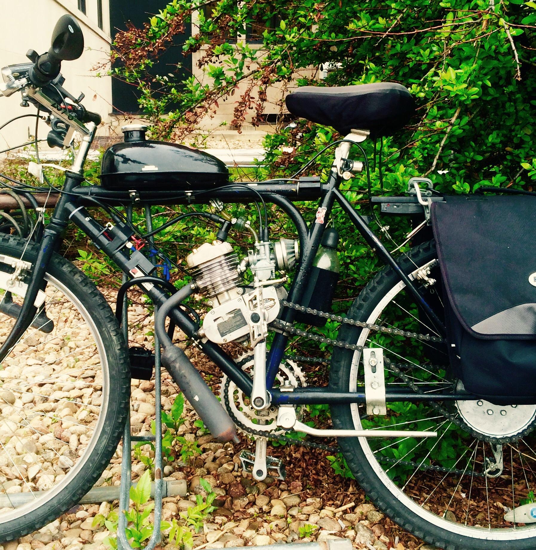 Yeti SB95: A black bicycle with a motorized engine attached, parked alongside greenery and gravel. The bike features a fuel tank on top, various cables, and a bike bag mounted on the rear.