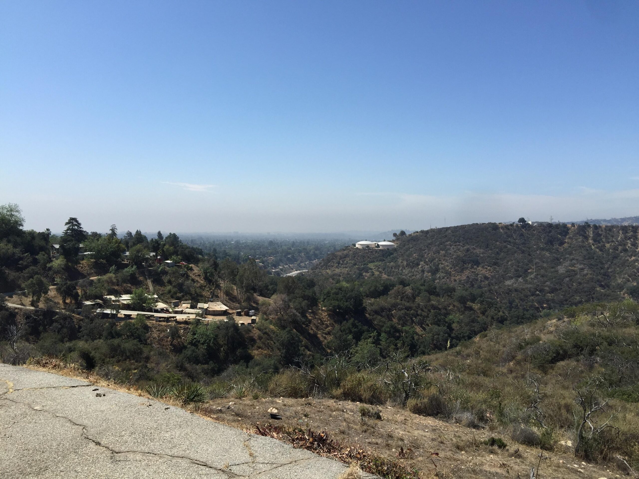A scenic view of rolling hills and valleys under a clear blue sky, with a sparse landscape featuring scattered trees and patches of vegetation. In the foreground, a winding road leads down into a valley where a few structures are visible. The distant horizon is slightly obscured by haze. El Prieto mountain bike trail.