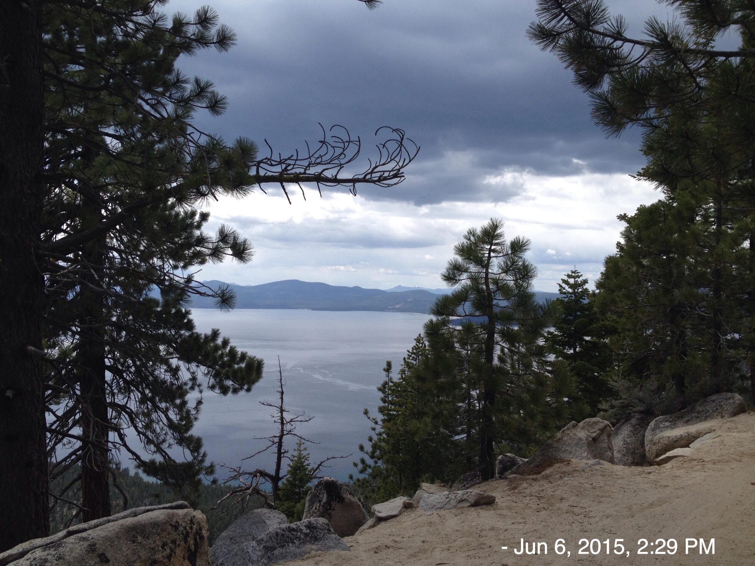 A scenic view of a lake surrounded by mountains, framed by pine trees. The sky is partially cloudy, creating a tranquil atmosphere. In the foreground, a sandy path and rocky terrain are visible, leading to the serene water in the background. The image is timestamped June 6, 2015, at 2:29 PM. Tahoe Rim Trail: Tahoe Meadows to Tunnel Creek Road / Flume Trail mountain bike trail.