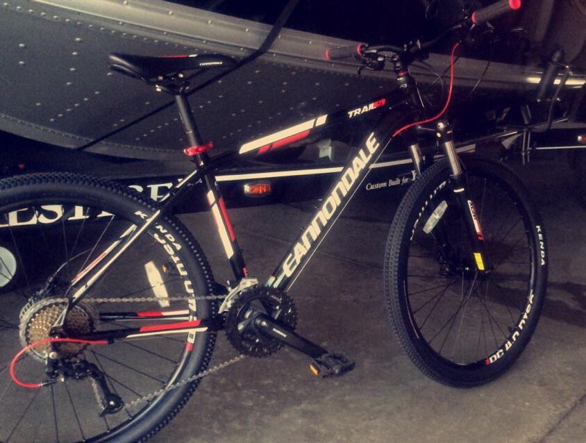 Cannondale Trail 5: A black Cannondale mountain bike with red and white accents, parked on a concrete floor next to a silver surface that appears to be part of a vehicle. The bike features knobby tires, a visible chain, and a multi-speed gear system.