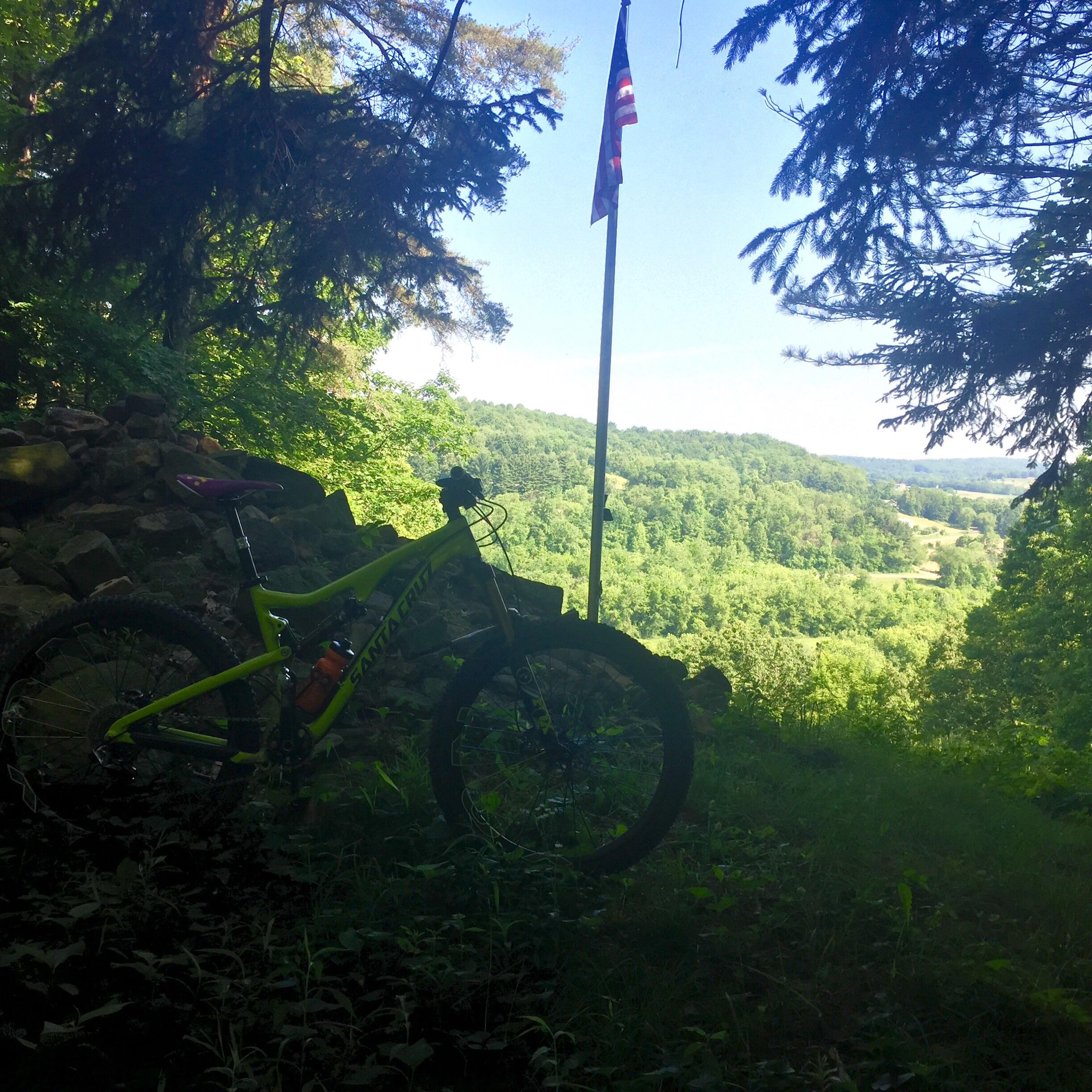 A vibrant green mountain bike rests beside a stone outcrop, with a tall American flag visible in the background against a scenic view of lush green hills and blue sky. Sunlight filters through the trees, creating a serene outdoor atmosphere. Camp Tuscazoar mountain bike trail.