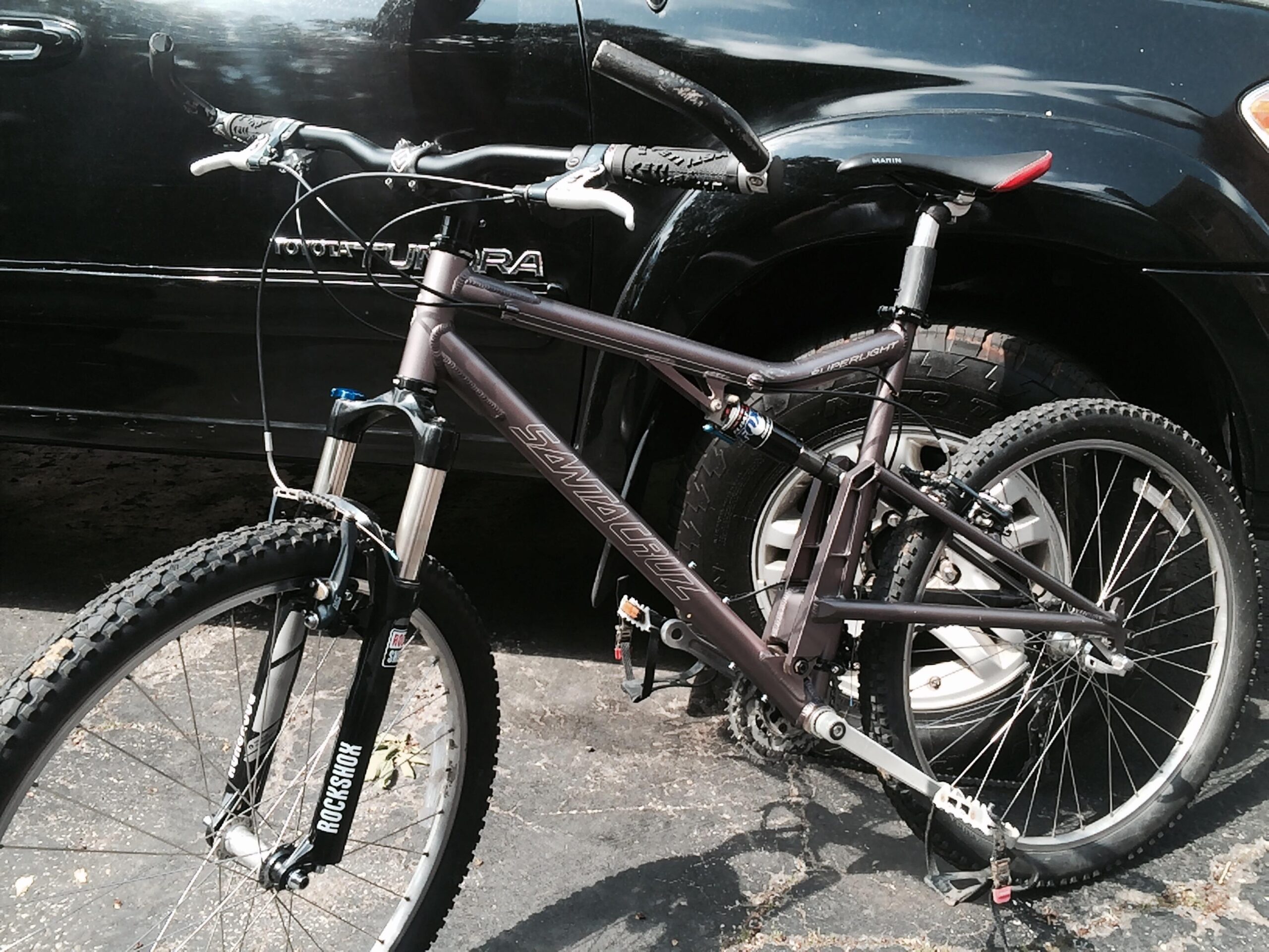 Santa Cruz Superlight: A mountain bike with a dark frame parked next to a black SUV. The bike features suspension forks, knobby tires, and a prominent brand logo on the fork. The SUV's wheel is partially visible in the background. The scene is set on a paved surface.