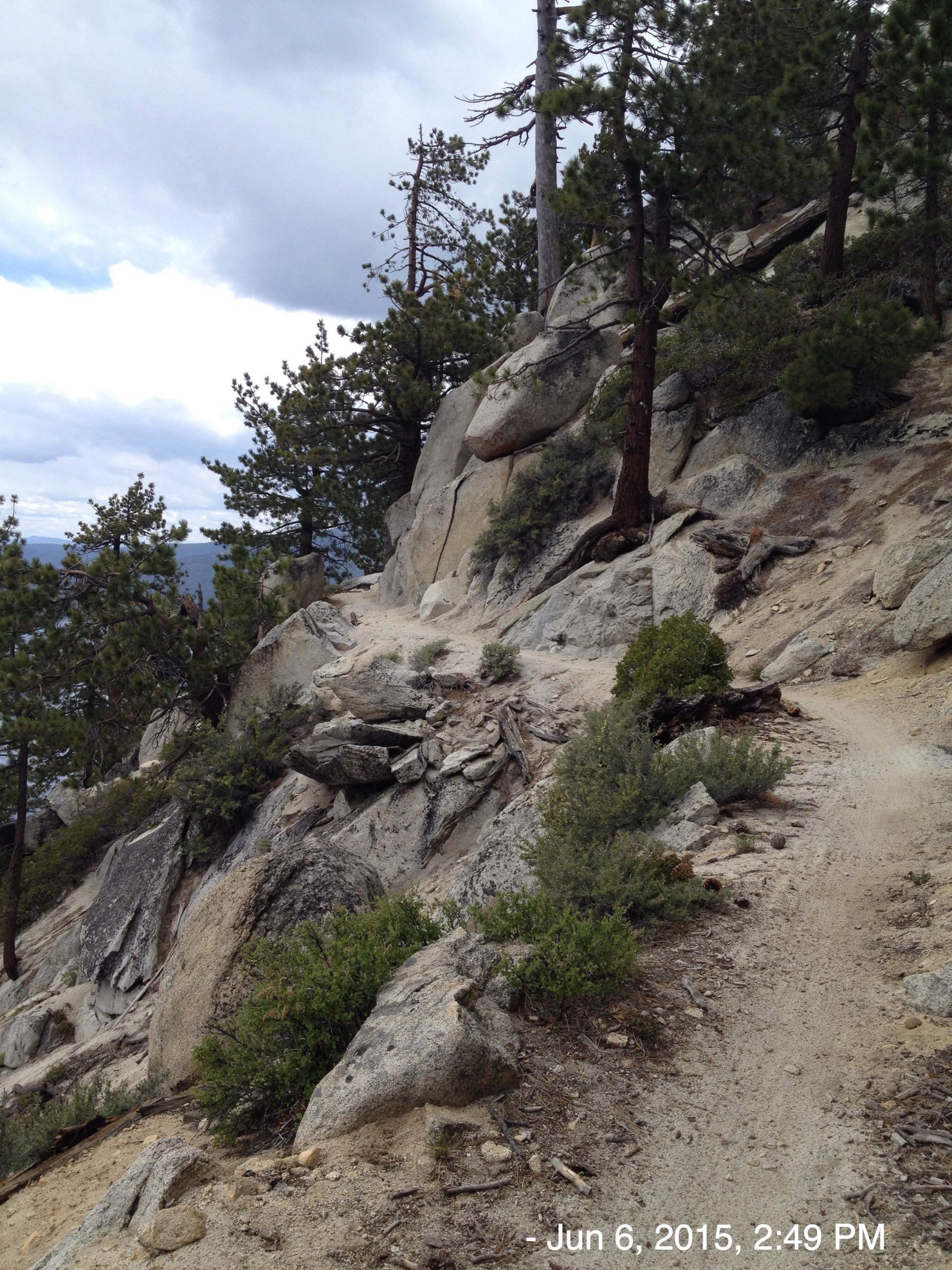 A rocky hiking trail winding through a mountainous terrain, with tall pine trees flanking the path. The sky is overcast, adding to the natural wilderness ambiance of the scene. Tahoe Rim Trail: Tahoe Meadows to Tunnel Creek Road / Flume Trail mountain bike trail.