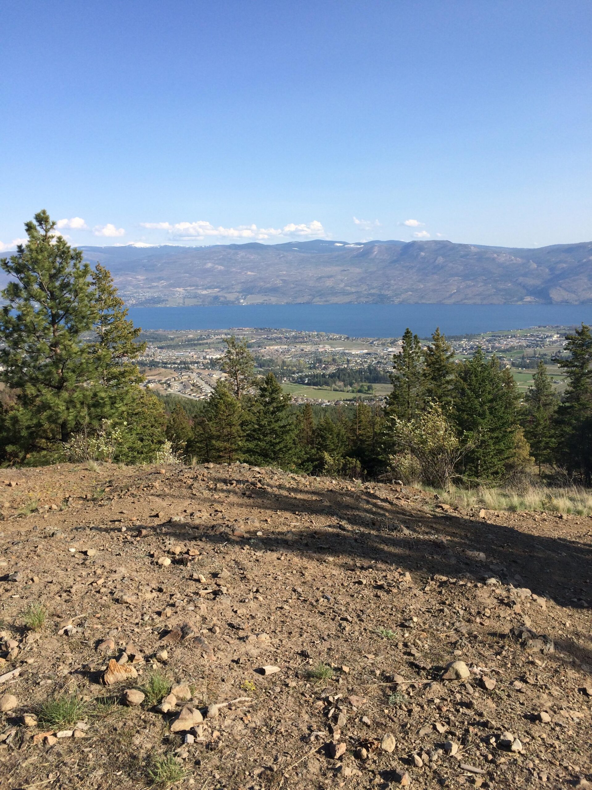 A panoramic view of a valley featuring a lake surrounded by mountains, captured from a rocky, elevated landscape. The foreground shows sparse vegetation and scattered rocks, while trees can be seen in the midground. The sky is clear with a few clouds, allowing for a bright and sunny atmosphere. Smith Creek mountain bike trail.