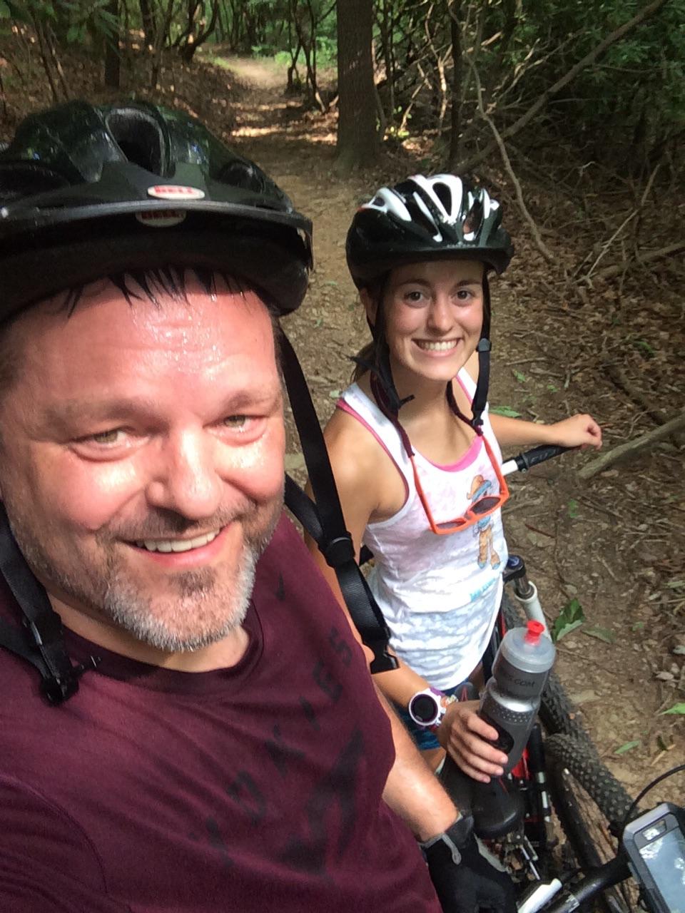 A man and a woman smiling while taking a selfie on a biking trail. Both are wearing helmets and have bikes alongside them. The background features lush greenery and a winding dirt path. The woman holds a water bottle and is wearing a tank top, while the man has a damp shirt from exertion. Woolwine Trails [Shiners Revenge] mountain bike trail.