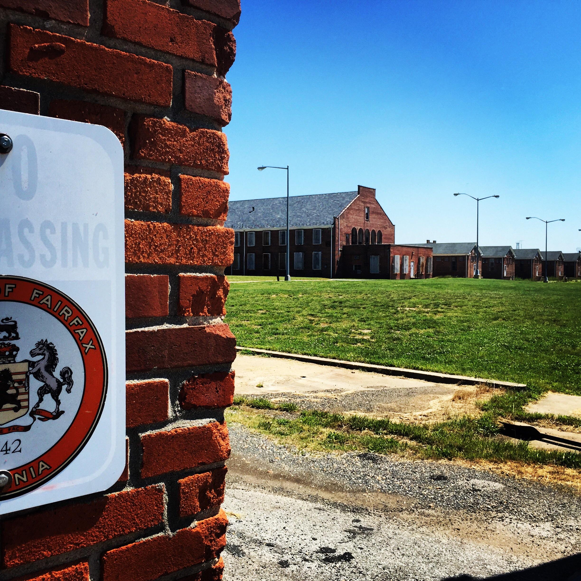 An image showing a brick wall with a "No Trespassing" sign attached, framing a view of an expansive green lawn and several old brick buildings in the background under a clear blue sky. The scene conveys an abandoned or restricted area. Laurel Hill Park mountain bike trail.