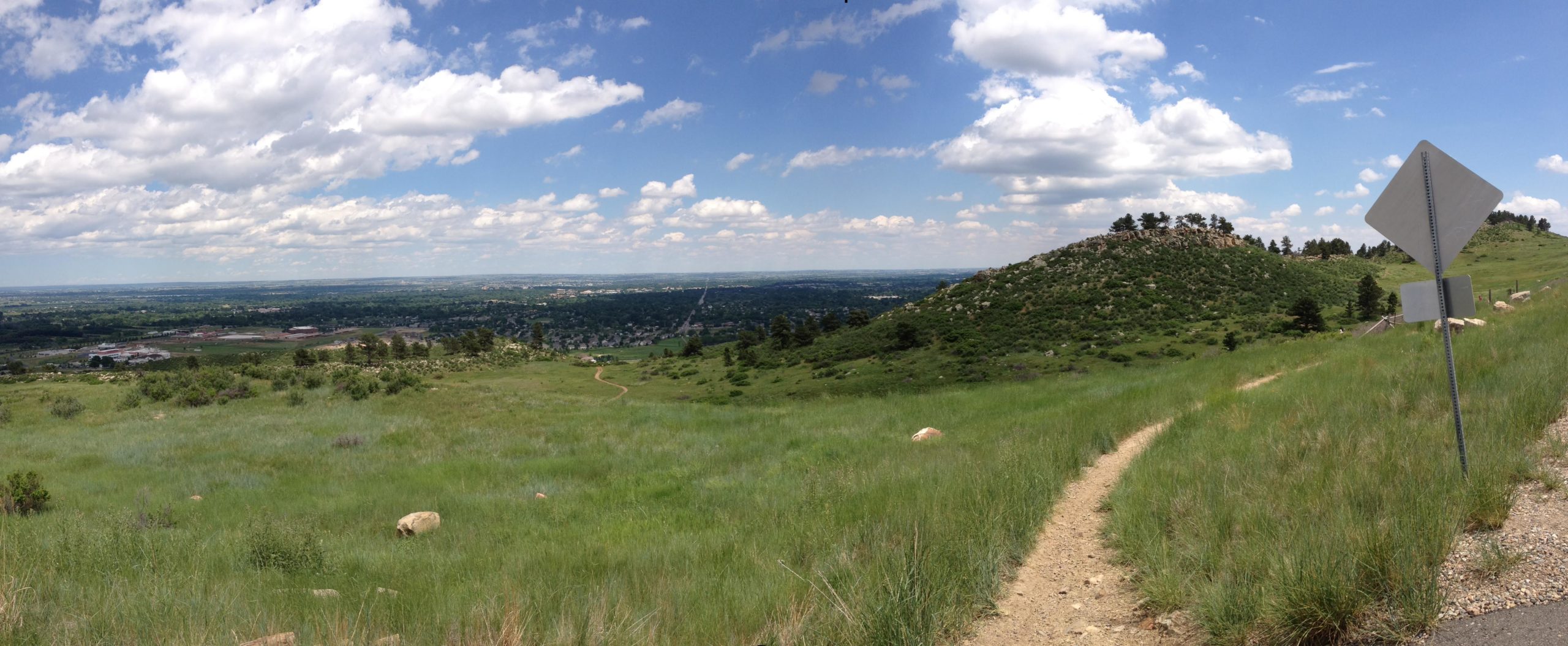A panoramic view of a rolling landscape featuring lush green hills and a winding dirt path. In the background, a small rocky hill is visible under a bright blue sky adorned with fluffy white clouds. There are hints of urban development in the valley below, with a few buildings and roads peeking through the greenery. A traffic sign is partially visible on the right side of the image. Maxwell's mountain bike trail.