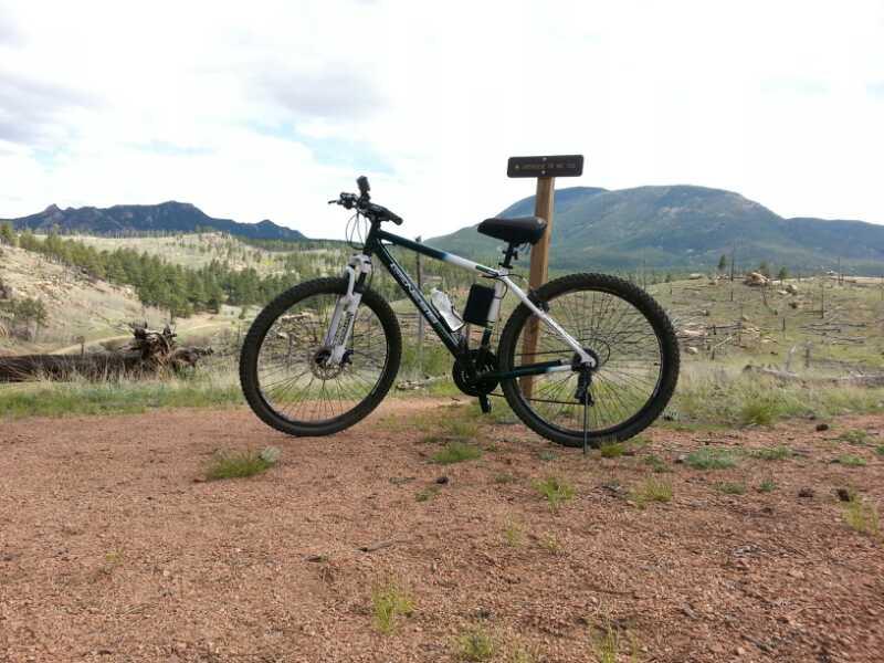 A mountain bike parked on a gravel path with a wooden sign in the background, set against a backdrop of rolling hills and mountains under a cloudy sky. The landscape includes patches of grass and scattered trees, indicating a natural outdoor environment. Buffalo Creek mountain bike trail.
