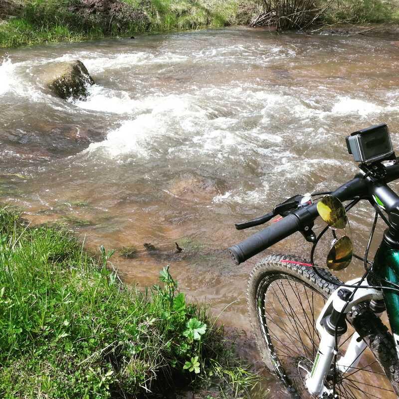A mountain bike leaning against a grassy bank near a flowing stream. The water is bubbling over rocks, with sunlight reflecting off the surface. The bike features a camera mounted on the handlebars and sunglasses resting on the grips. Lush green vegetation surrounds the scene, creating a peaceful outdoor atmosphere. Buffalo Creek mountain bike trail.