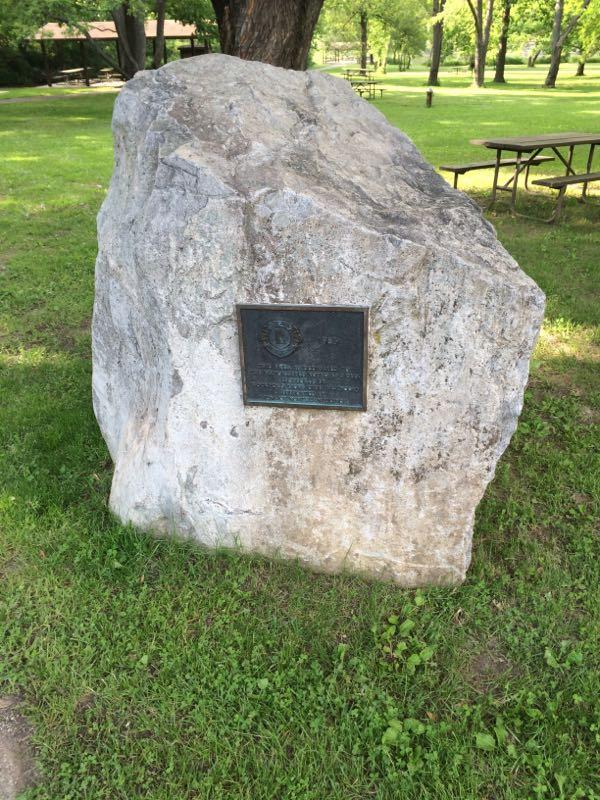 A large natural rock with a bronze plaque mounted on its side, set on a grassy area in a park. In the background, there are trees and picnic tables. Rock Cut State Park mountain bike trail.