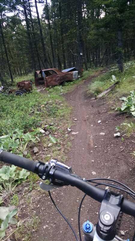 A view from the handlebars of a mountain bike riding along a dirt trail, surrounded by greenery and trees. In the background, there are two rusted, abandoned vehicles partially hidden by vegetation. Smith Creek mountain bike trail.