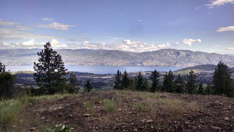 A panoramic view of a lake surrounded by mountains, with trees in the foreground and a partly cloudy sky. The landscape features a mix of greenery and rocky terrain, showcasing the natural beauty of the area. Smith Creek mountain bike trail.
