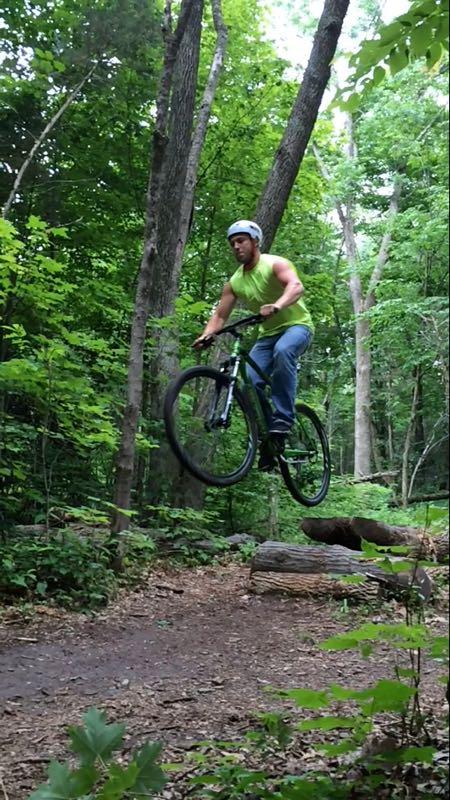 A cyclist in a green tank top and helmet performing a jump over a log on a mountain bike in a wooded area. Surrounding trees and foliage create a lush, green background, emphasizing the outdoor adventure. Nockamixon State Park mountain bike trail.