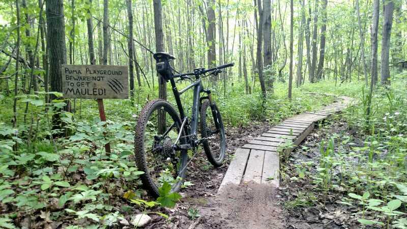 A mountain bike parked beside a wooden sign that reads "Puma Playground: Beware not to get mauled!" in a forested area. The bike is positioned near a narrow wooden trail leading through lush greenery and trees. Hickory Ridge mountain bike trail.