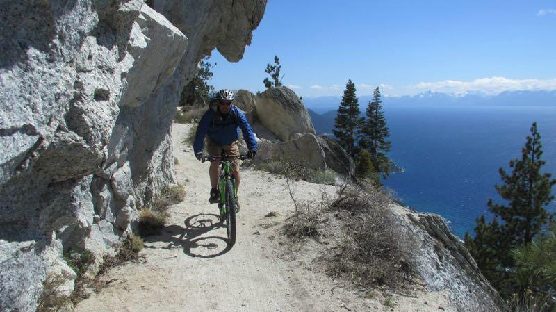 A mountain biker riding along a narrow dirt trail surrounded by rocky cliffs and trees, with a scenic view of a lake and mountains in the background under a clear blue sky. Flume Trail mountain bike trail.