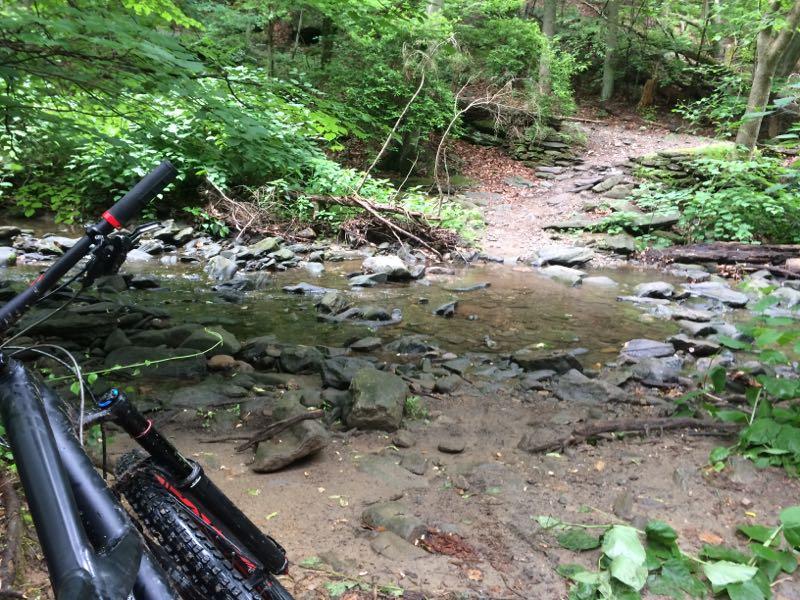 A mountain bike resting on the ground next to a clear, shallow stream surrounded by lush green foliage and rocky terrain, leading into a trail in a wooded area. Wissahickon Valley Park mountain bike trail.