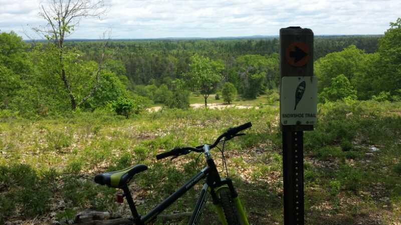 A mountain bike resting beside a trail marker on a grassy overlook with a panoramic view of green trees and valleys under a cloudy sky. The marker indicates the direction of the nearby trail. Hanson Hills mountain bike trail.