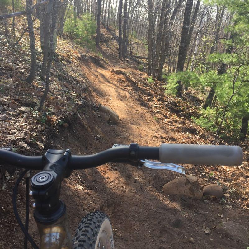 A view of a mountain bike handlebar in the foreground, with a dirt trail winding through a wooded area in the background. The trail is surrounded by trees and has patches of sunlight filtering through the leaves. Mt Ashwabay Singletrack mountain bike trail.